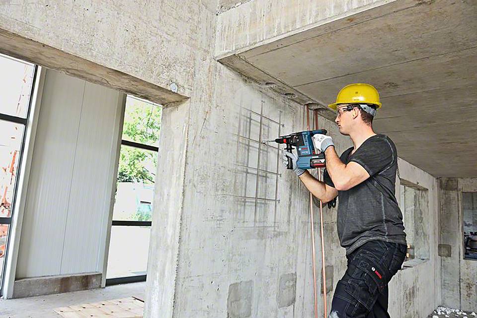Ein Bauarbeiter mit gelbem Helm bohrt mit einer elektrischen Bohrmaschine in eine Betonwand auf einer Baustelle.
