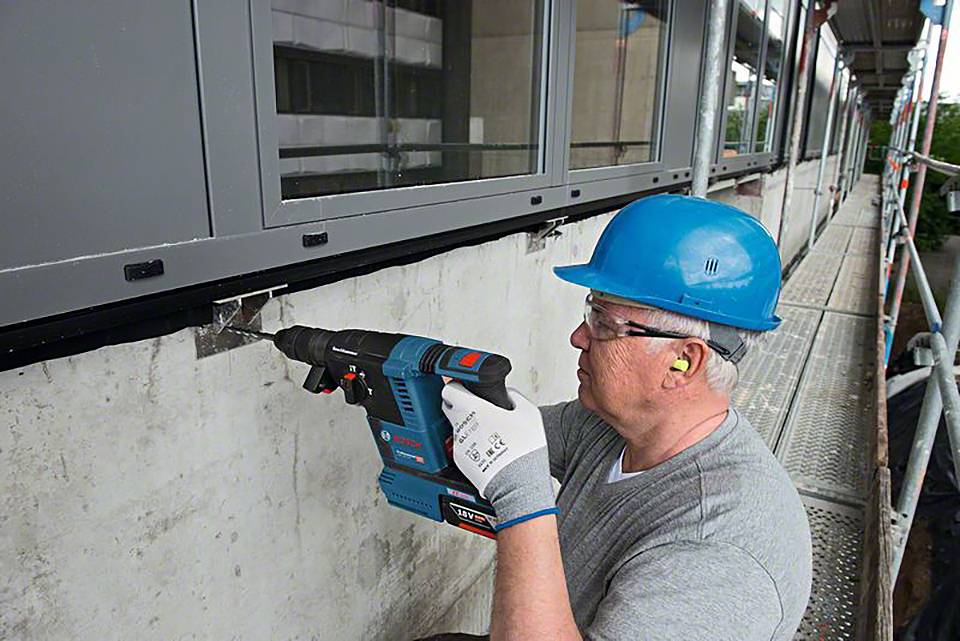 Ein Arbeiter in Sicherheitsausrüstung benutzt eine Bohrmaschine, um eine Befestigung an einem Betonfundament unter Fenstern anzubringen.