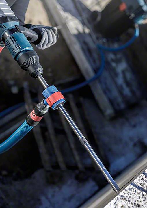 Ein Handwerker verwendet eine elektrische Bohrmaschine auf einer Baustelle. Im Hintergrund sind Bauwerkzeuge und Boden zu sehen.