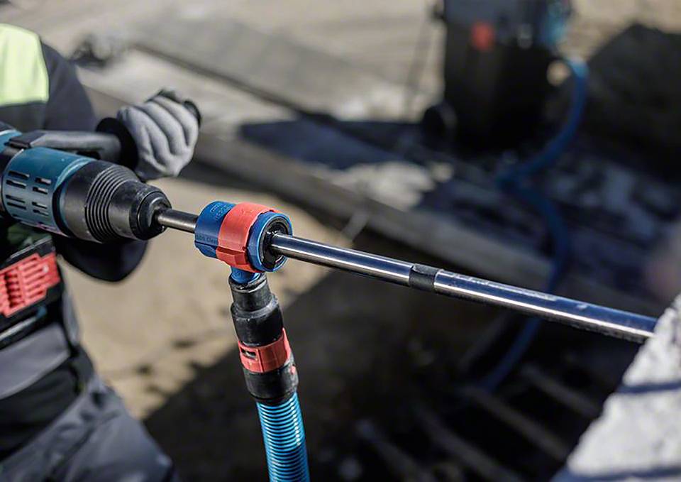 Arbeiter benutzt einen Bohrer mit blau-rotem Aufsatz auf einer Baustelle. Der Bohrer wird in Beton eingesetzt.