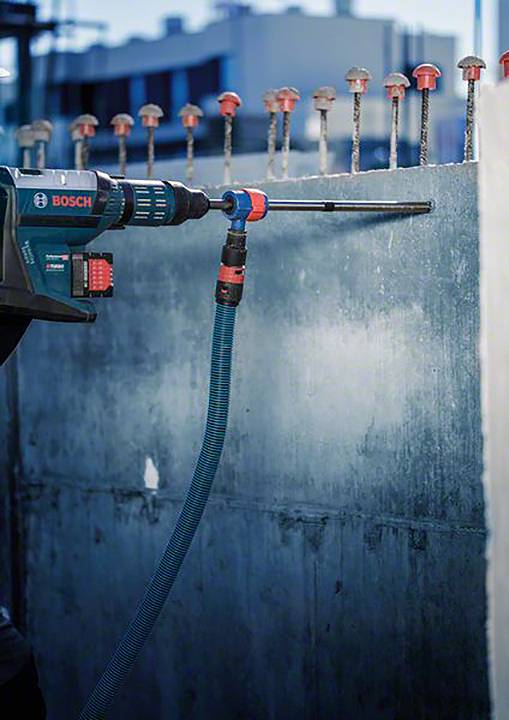 Eine Bohrmaschine wird benutzt, um in eine Betonwand mit vielen herausragenden Metallstangen zu bohren. Im Hintergrund sind Gebäude erkennbar.