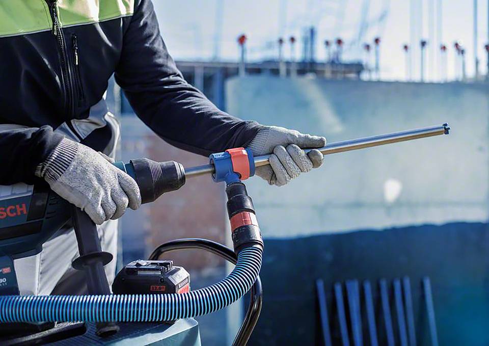 Eine Person mit Schutzkleidung hält eine Bohrmaschine vor einer unscharfen Baustellenkulisse. Sie arbeitet präzise mit dem Gerät.