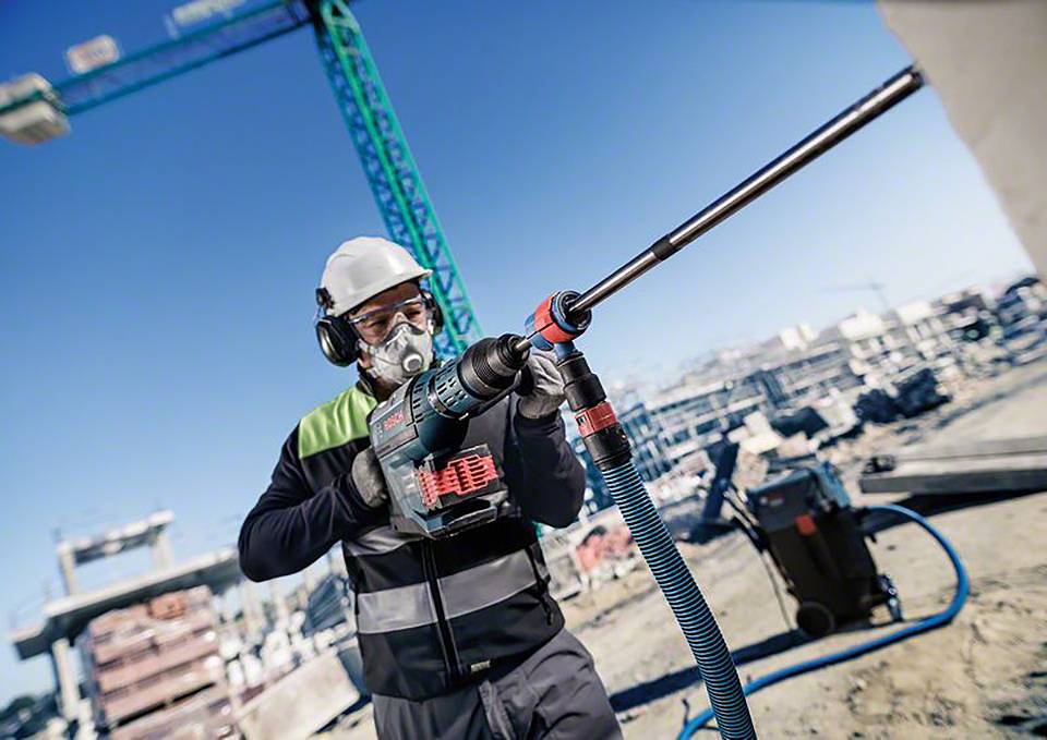 Ein Bauarbeiter mit Schutzbrille und Helm benutzt eine Bohrmaschine auf einer Baustelle. Im Hintergrund sind Kräne und Bauwerke.
