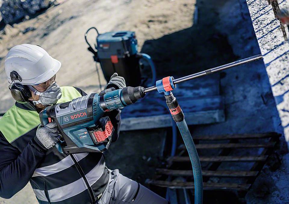 Eine Person mit Schutzkleidung und -ausrüstung bohrt mit einer Bosch-Bohrmaschine in eine Betonmauer auf einer Baustelle.