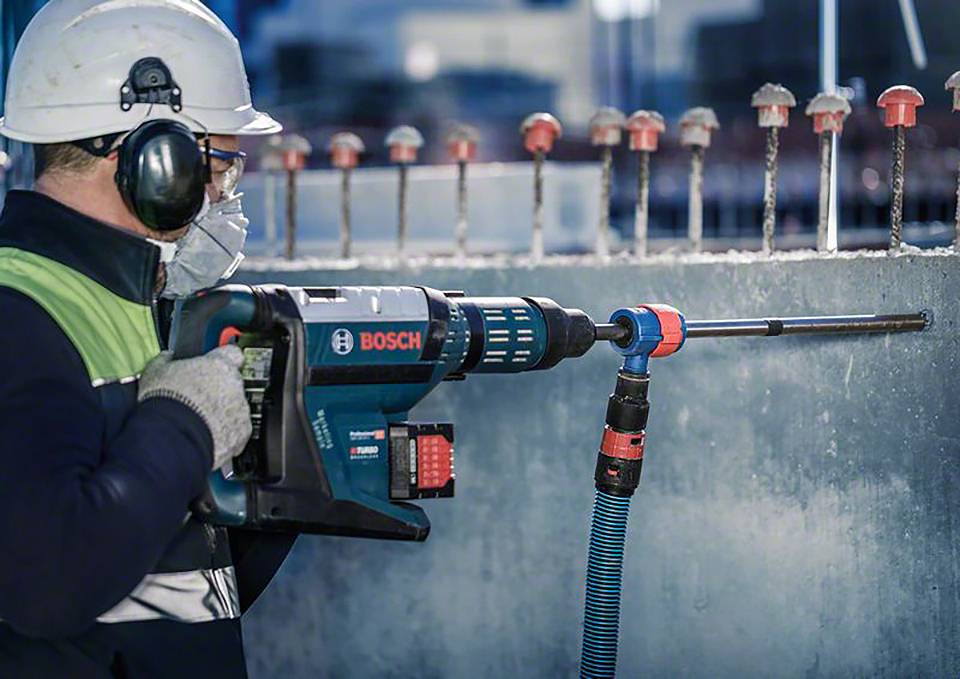 Ein Bauarbeiter mit Schutzhelm und Gehörschutz bohrt mit einer elektrischen Bohrmaschine ein Loch in eine Betonwand.