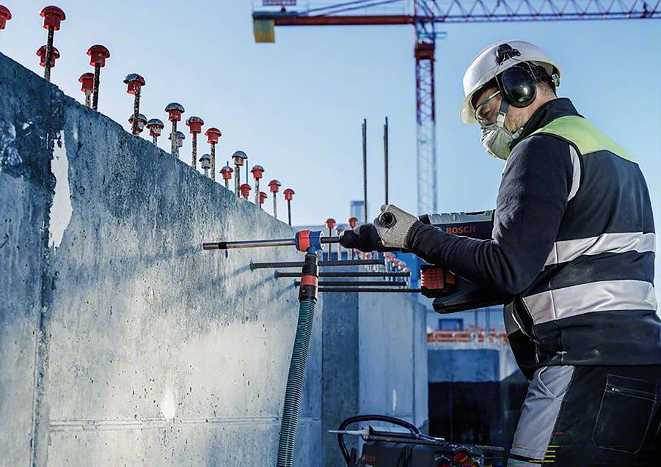 Bauarbeiter in Schutzkleidung bohrt mit Elektrowerkzeug in Betonwand. Kran im Hintergrund, Baustelle im Freien.