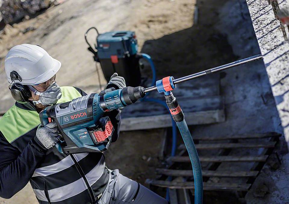 Arbeiter mit Sicherheitsausrüstung bohrt mit einem Bosch-Bohrhammer in eine Betonwand. Hintergrund zeigt Baustellenumgebung.