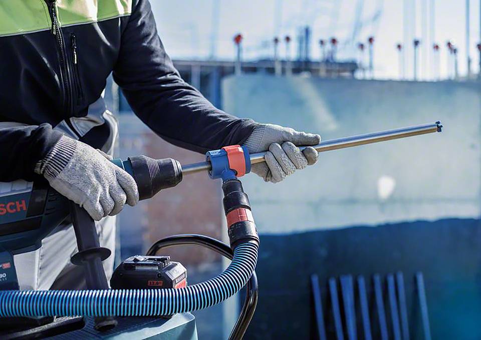 Eine Person mit Sicherheitsausrüstung arbeitet mit einer Bohrmaschine auf einer Baustelle. Im Hintergrund sind unscharfe Stahlträger zu sehen.