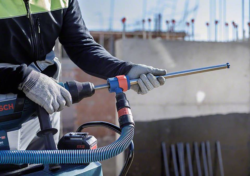 Eine Person im Bauarbeiter-Outfit verwendet ein Elektrowerkzeug mit einem langen Bohrer. Hintergrund zeigt eine unfertige Baustelle.