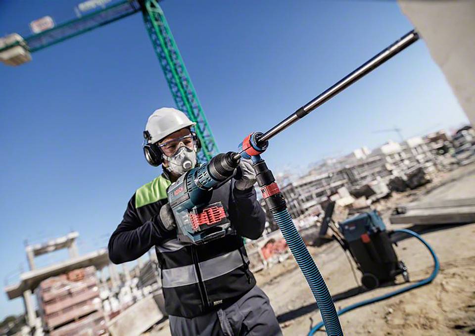 Ein Bauarbeiter mit Schutzbrille und Atemmaske benutzt eine elektrische Bohrmaschine auf einer Baustelle, im Hintergrund Kräne und Gebäude.