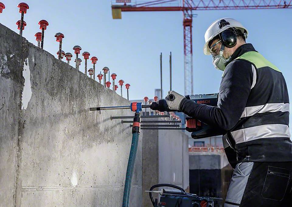 Ein Bauarbeiter trägt Schutzkleidung und bohrt ein Loch in eine Betonwand auf einer Baustelle, im Hintergrund ein Kran.