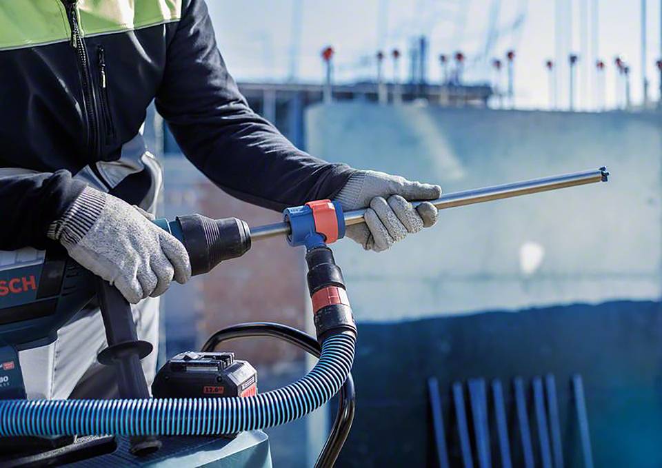 Arbeiter in Schutzkleidung nutzt ein Elektrowerkzeug mit Staubsaugeraufsatz auf einer Baustelle. Hintergrund zeigt unscharfe Strukturteile.