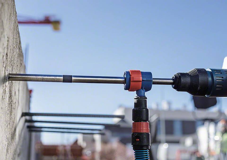 Ein Bohrhammer bearbeitet eine Betonwand. Im Hintergrund sind verschwommene Baugerüste und ein Kran sichtbar, was eine Baustellenszene andeutet.