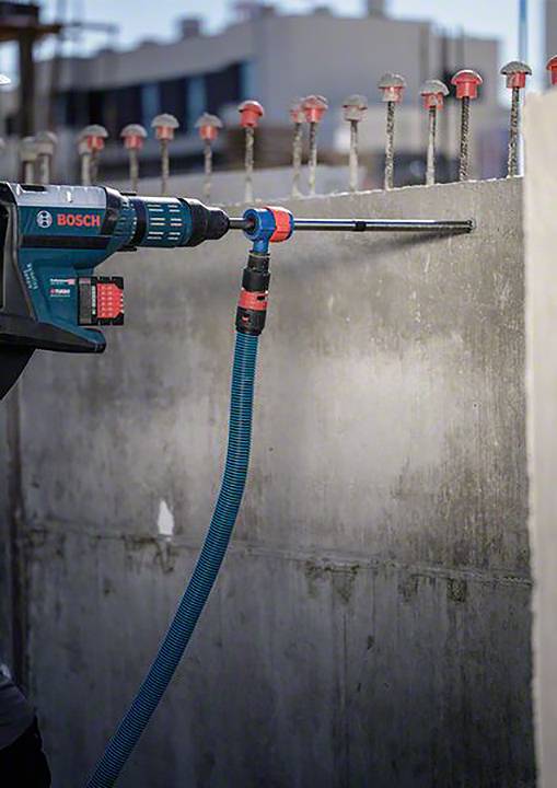 Eine blaue Bohrmaschine bohrt ein Loch in eine Betonwand. Oben sind herausragende Stahlstäbe zu sehen. Es ist eine Baustellenszene.