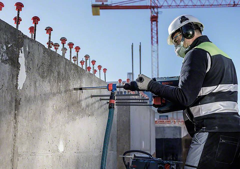 Ein Bauarbeiter bohrt mit einer Bohrmaschine in eine Betonwand. Im Hintergrund sind ein Kran und andere Baustellenstrukturen zu sehen.