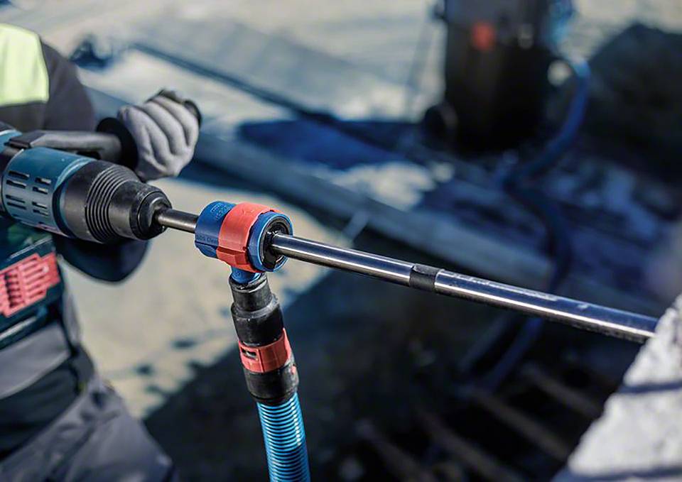 Arbeiter bohrt mit Elektrowerkzeug in Beton, Tragen von Schutzkleidung, Fokus auf Bohrkopf. Umgebung deutet Baustelle an.