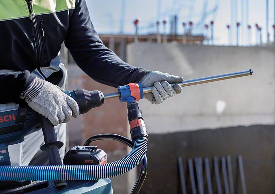 Eine Person hält eine Bohrmaschine mit einem langen Bohrer, trägt Handschuhe und Arbeitskleidung. Im Hintergrund eine Baustelle.