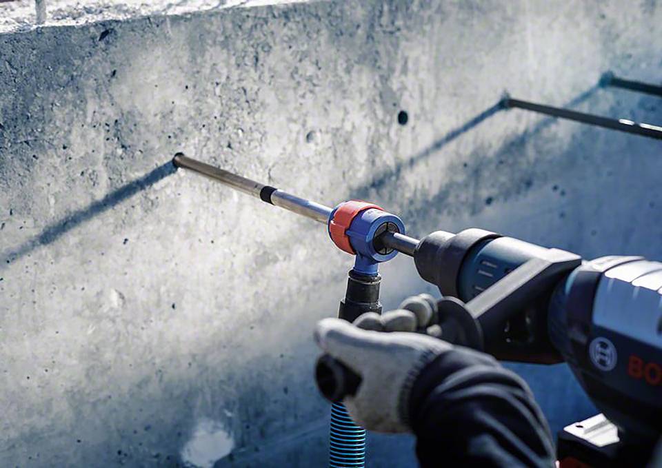 Eine Person bohrt mit einem Bohrhammer ein Loch in eine Betonwand. Der Bohrer hat ein langes Bit und erzeugt Staub.