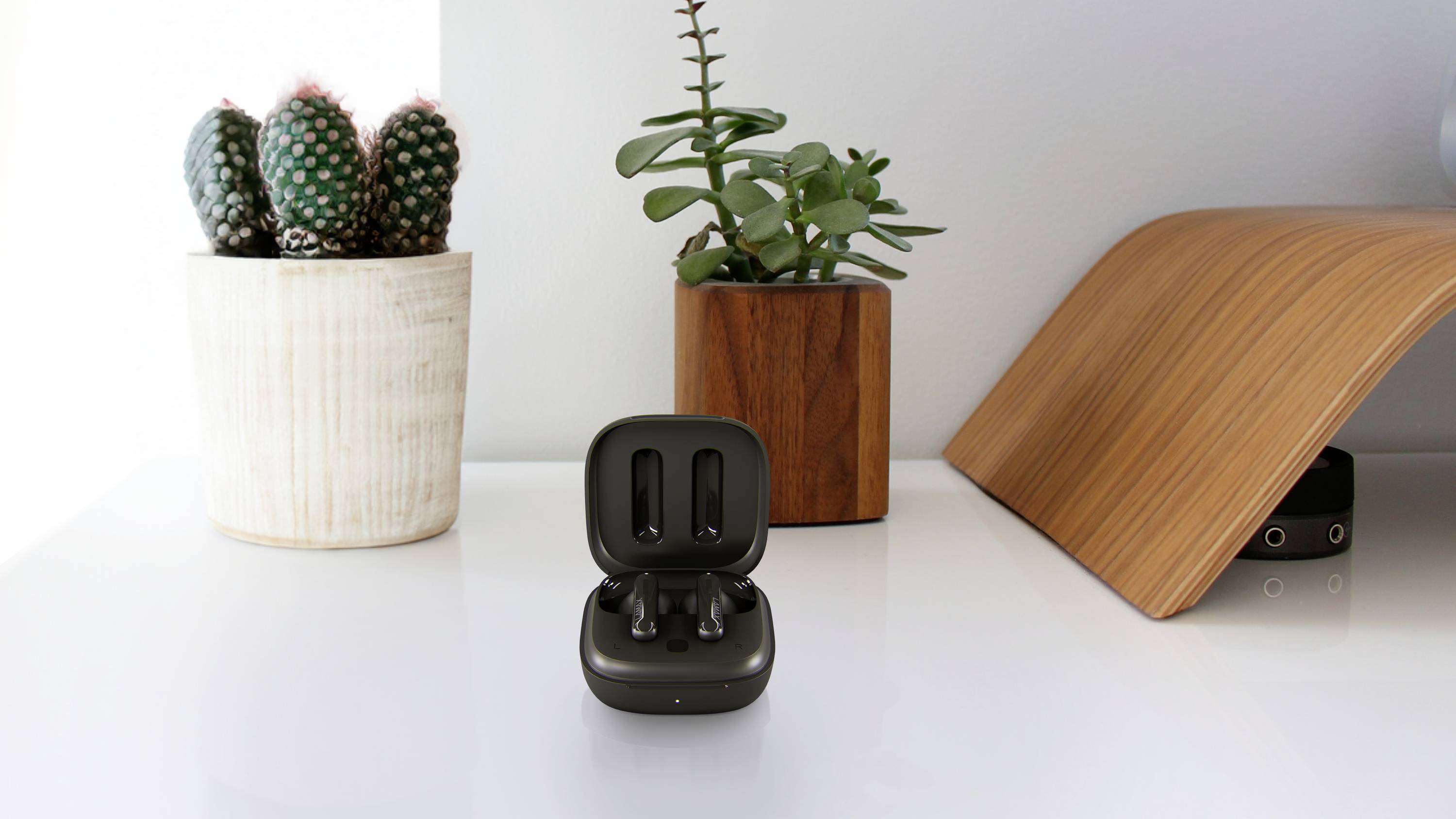 Ein schwarzes kabelloses Ohrhörer-Etui steht geöffnet auf einem weißen Tisch, umgeben von zwei Blumentöpfen und einem Holz-Dekorstück.