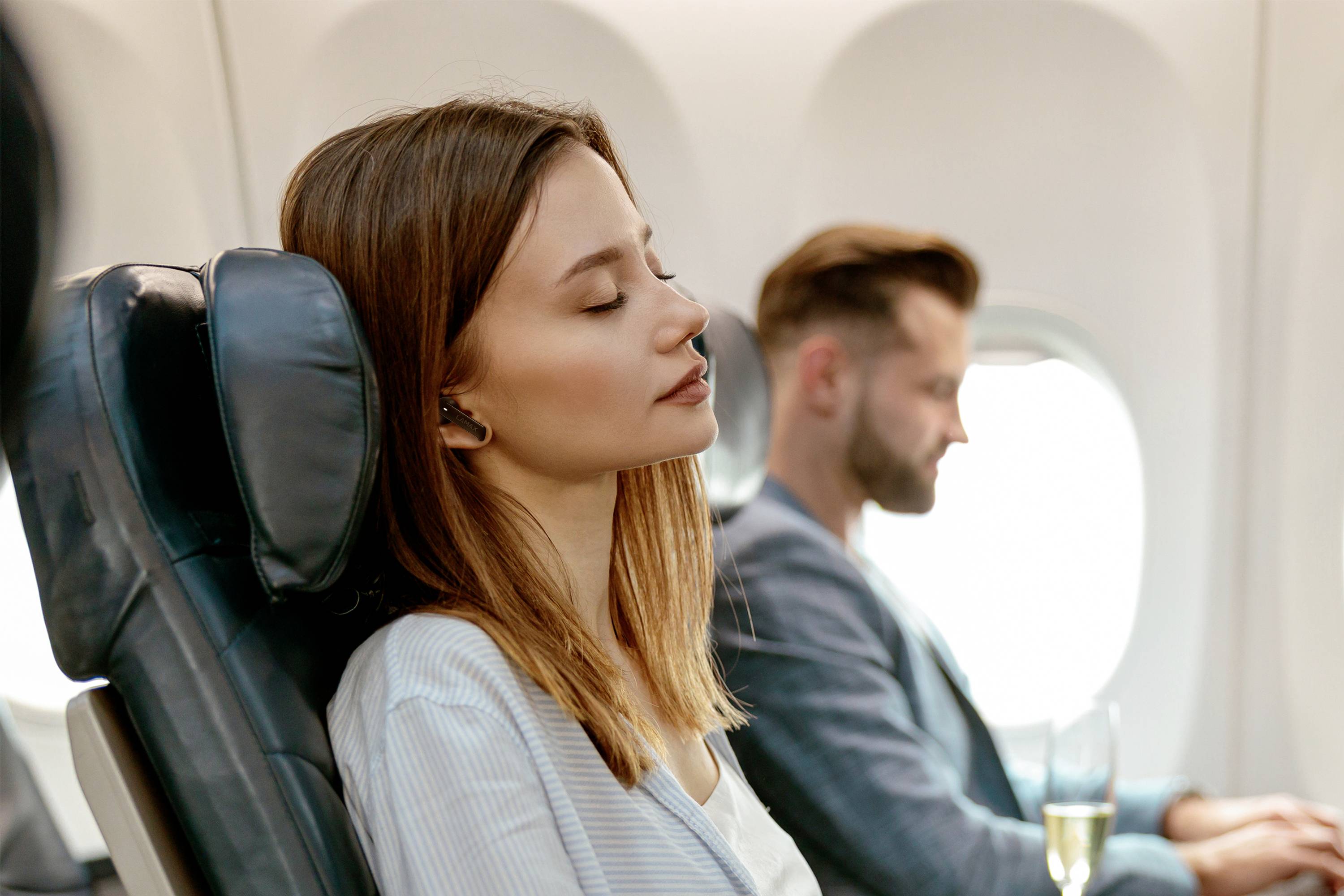 Eine Frau sitzt entspannt in einem Flugzeugsitz, Augen geschlossen, mit Kopfhörern. Im Hintergrund sitzt ein Mann, der aus dem Fenster schaut.