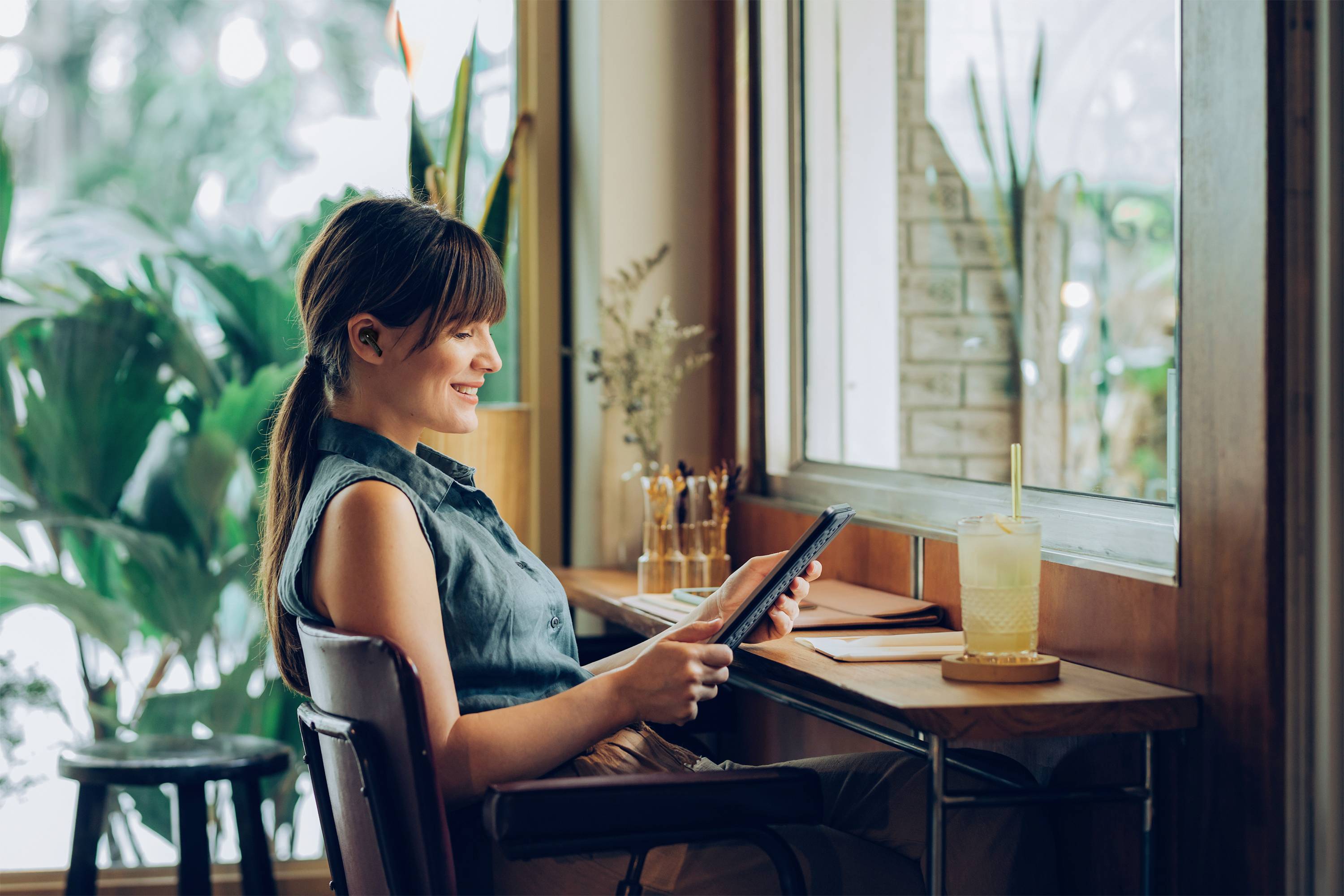 Frau sitzt an Tisch in Café, schaut auf Tablet und lächelt. Im Hintergrund Pflanzen und Fenster.
