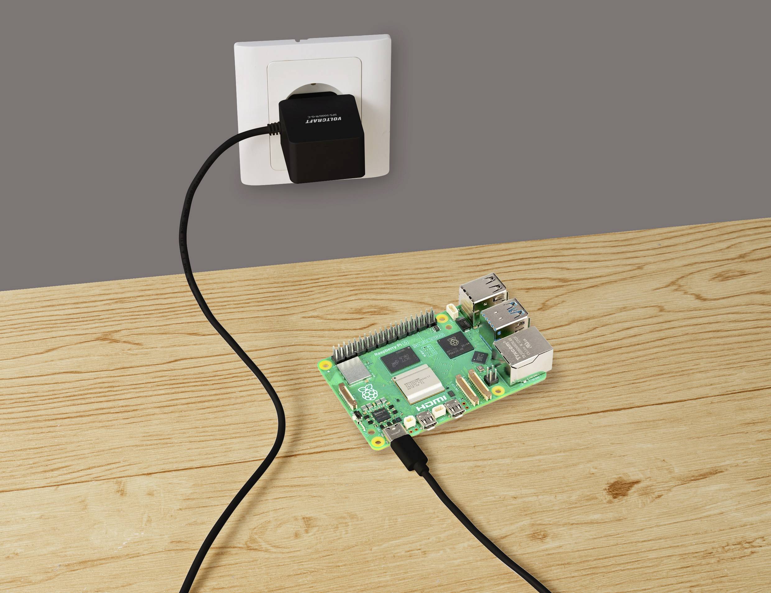 'Raspberry Pi' Computerplatine auf einem Holztisch, verbunden mit einem Stromadapter in einer Wandsteckdose.