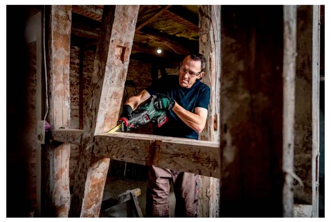 „Eine Person, die eine Kreissäge verwendet, um Holz an einem Baustellenstandort zu schneiden, trägt Schutzbrille und Handschuhe und ist von Holzbalken und Werkzeugen umgeben.