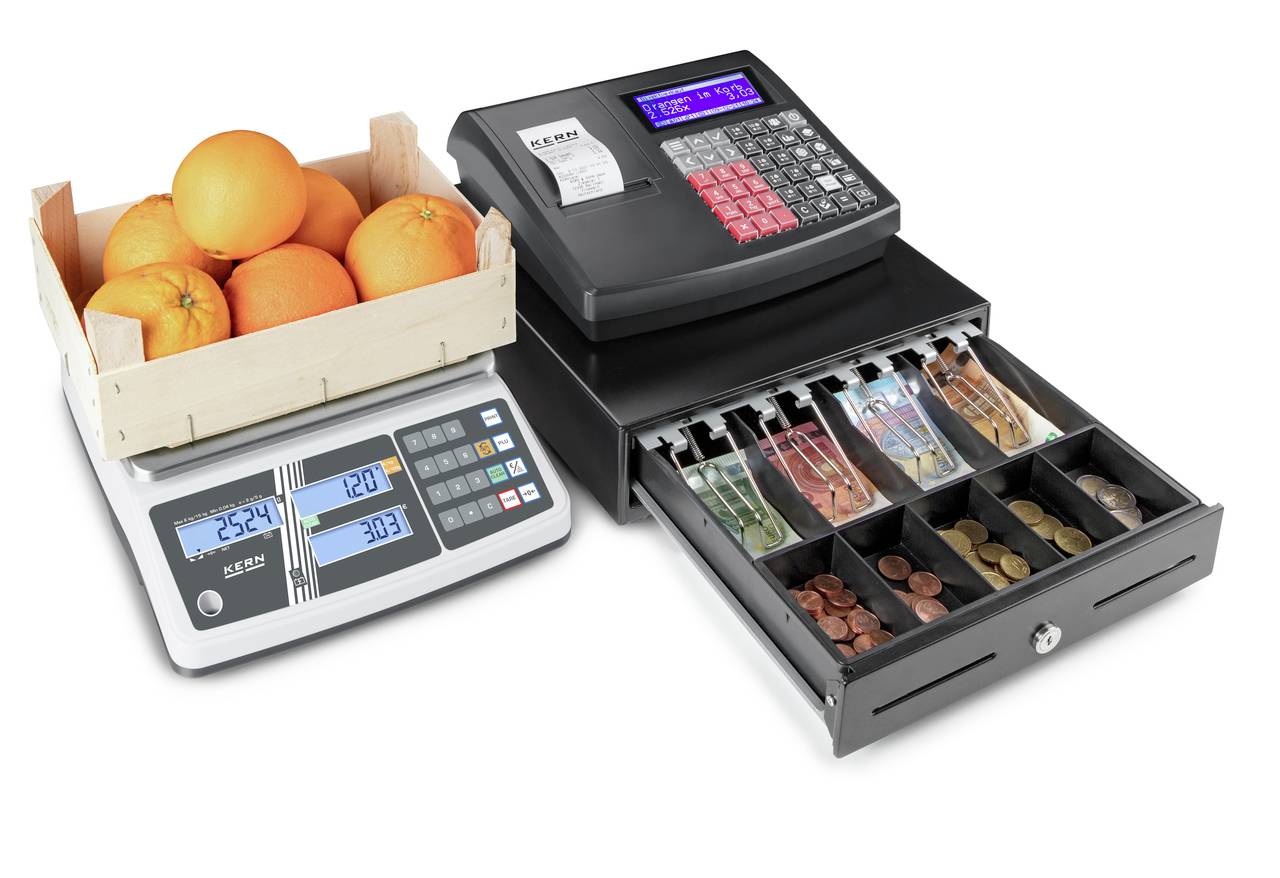 Digitale Obstwaage mit Holzkorb voller Orangen neben offener Kassenschublade mit sortiertem Bargeld und Registrierkasse.