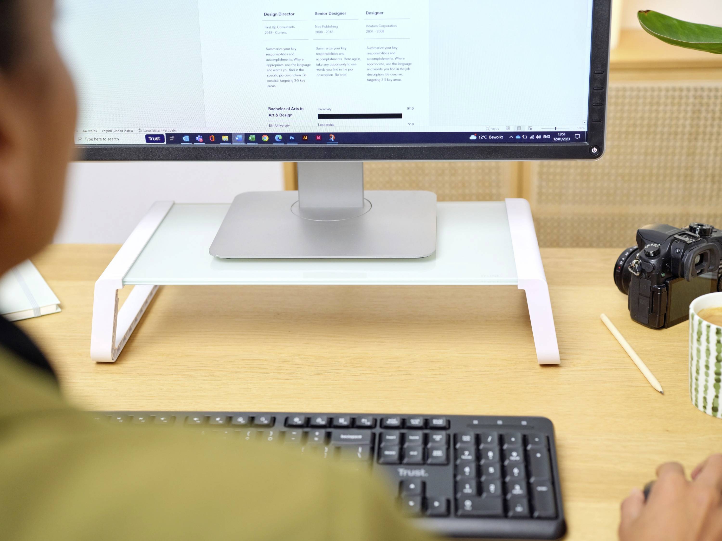 Person sitzt vor einem Schreibtisch, betrachtet einen Monitor mit Text. Auf dem Tisch befinden sich eine Tastatur, Kamera und Tasse.
