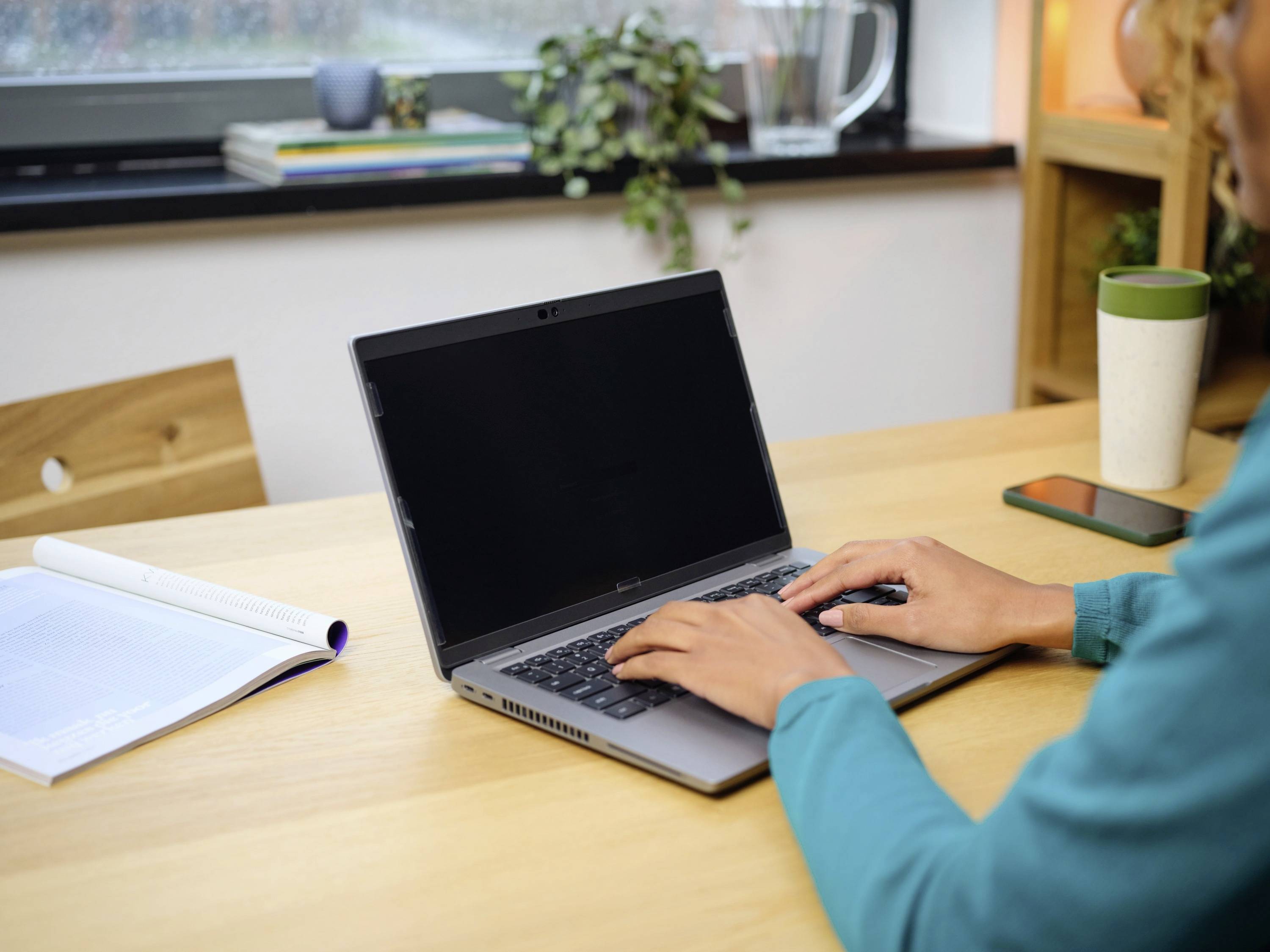 Person arbeitet an einem Laptop auf einem Holztisch. Daneben liegen ein geöffnetes Buch und ein Smartphone. Im Hintergrund sind Pflanzen sichtbar.