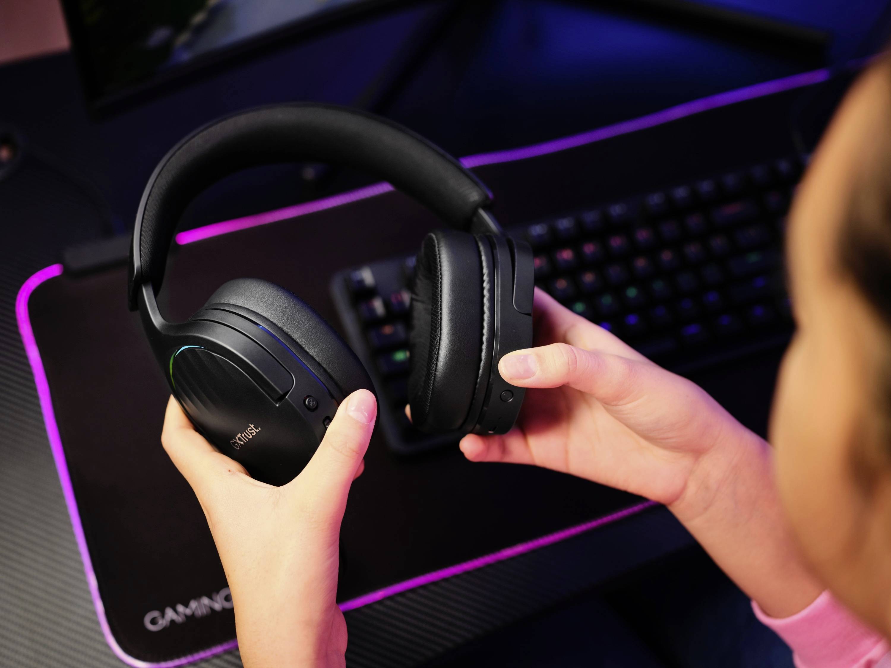 Eine Person hält ein schwarzes Headset vor einem beleuchteten Desktop-Computer mit Tastatur. Die Szene deutet auf eine Spielsituation hin.