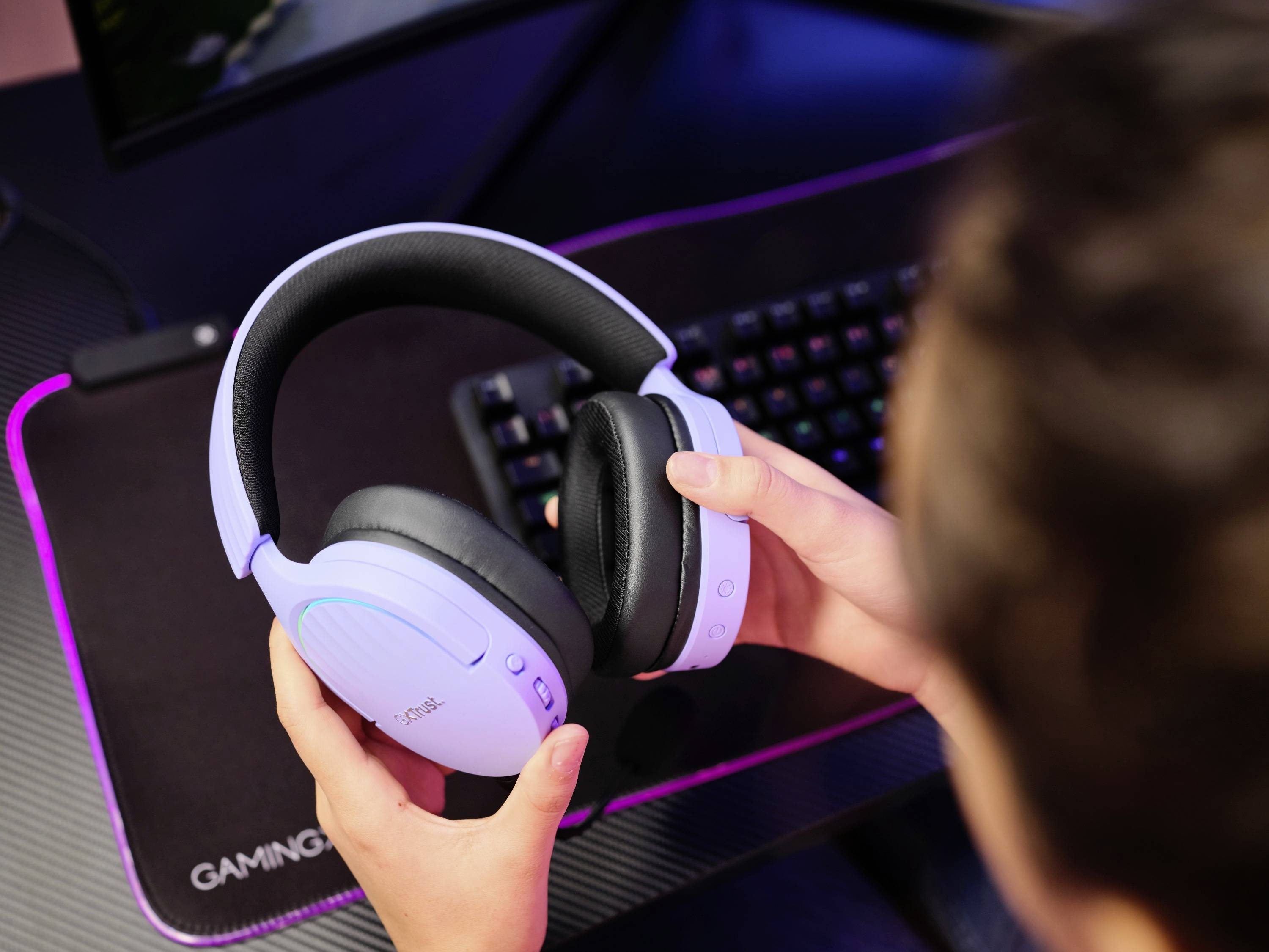 Eine Person hält ein kabelloses Headset über einem Schreibtisch mit Mauspad und beleuchteter Tastatur.