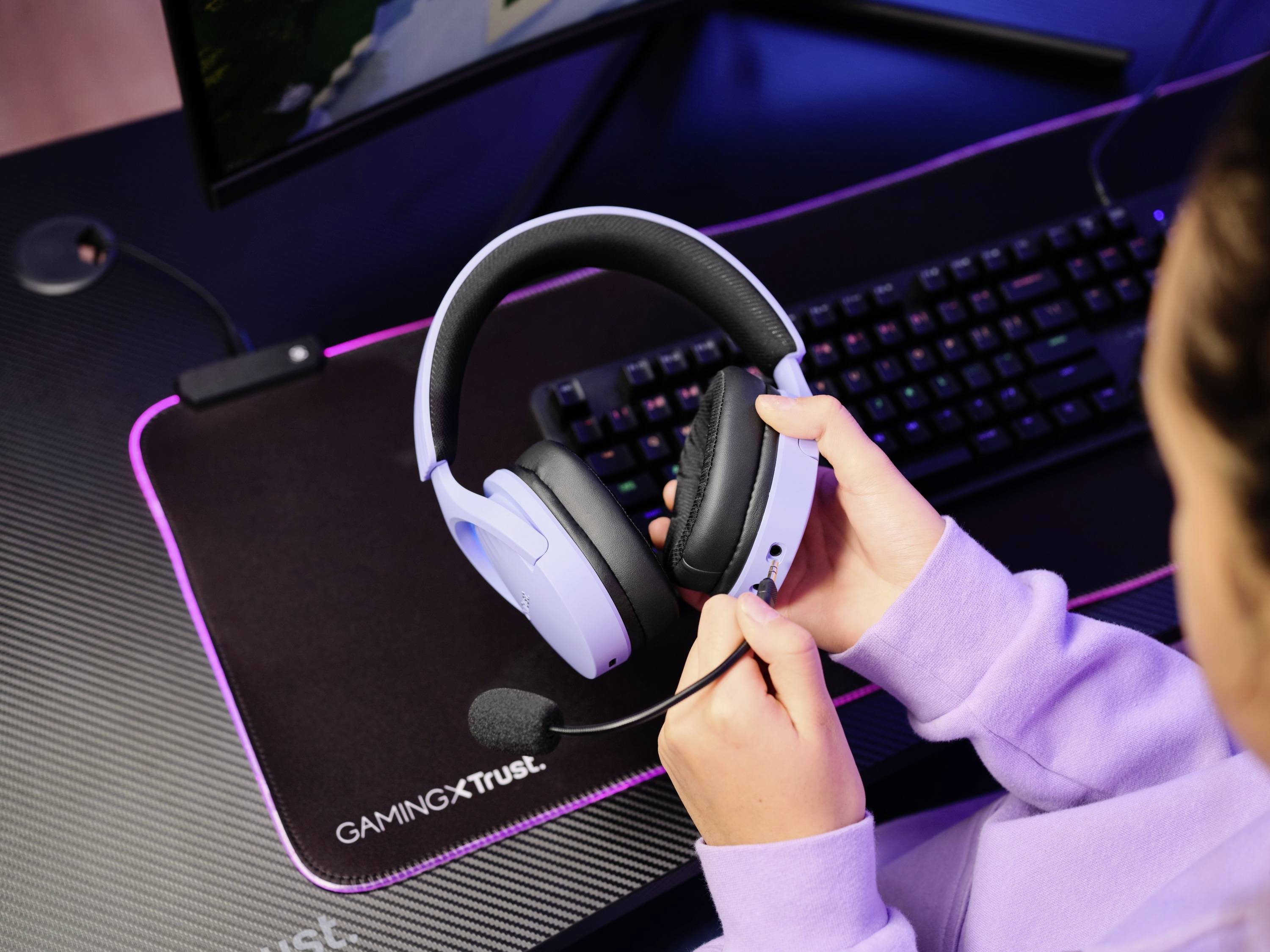Eine Person hält ein weißes Gaming-Headset über einem Computer mit sichtbarer Tastatur und Mauspad auf einem Tisch.