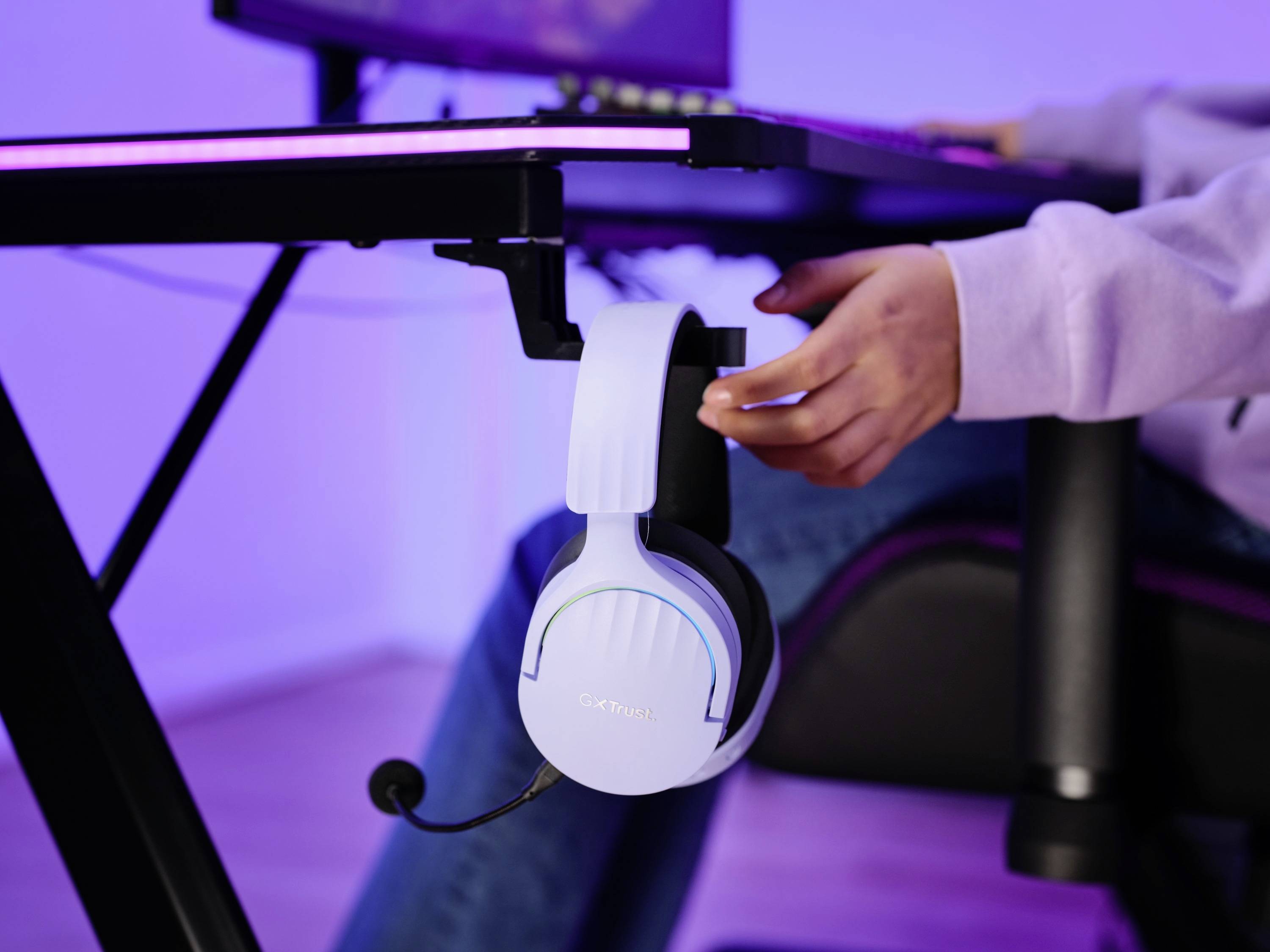 Eine Person hängt ein weißes Headset an einen Tischhaken. Es scheint ein Gaming-Setup mit lilafarbener Beleuchtung zu sein.