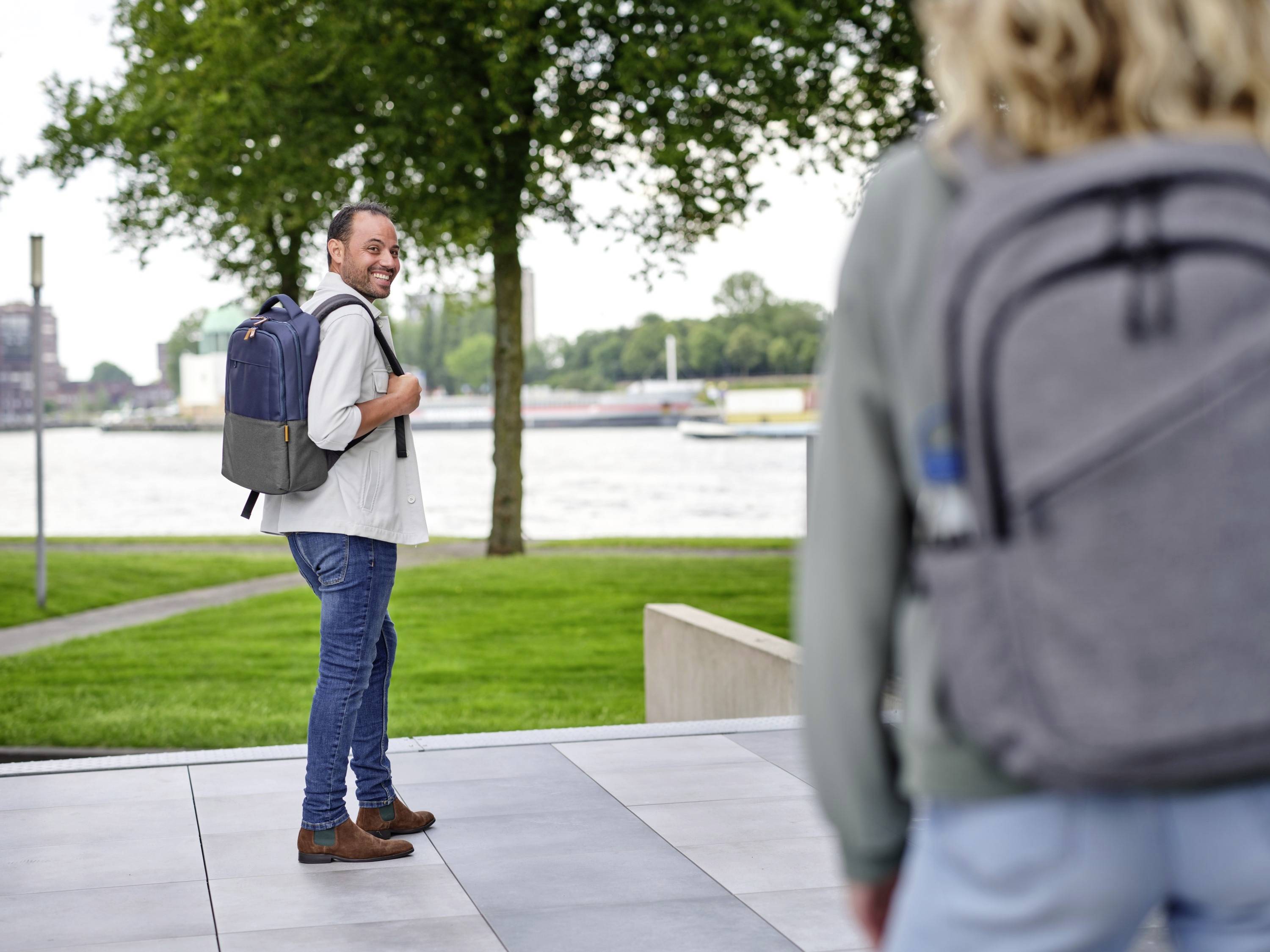 Ein Mann mit Rucksack schaut lächelnd zurück, während er in einem Park mit Blick auf einen Fluss spaziert. Im Vordergrund unscharfe Person.