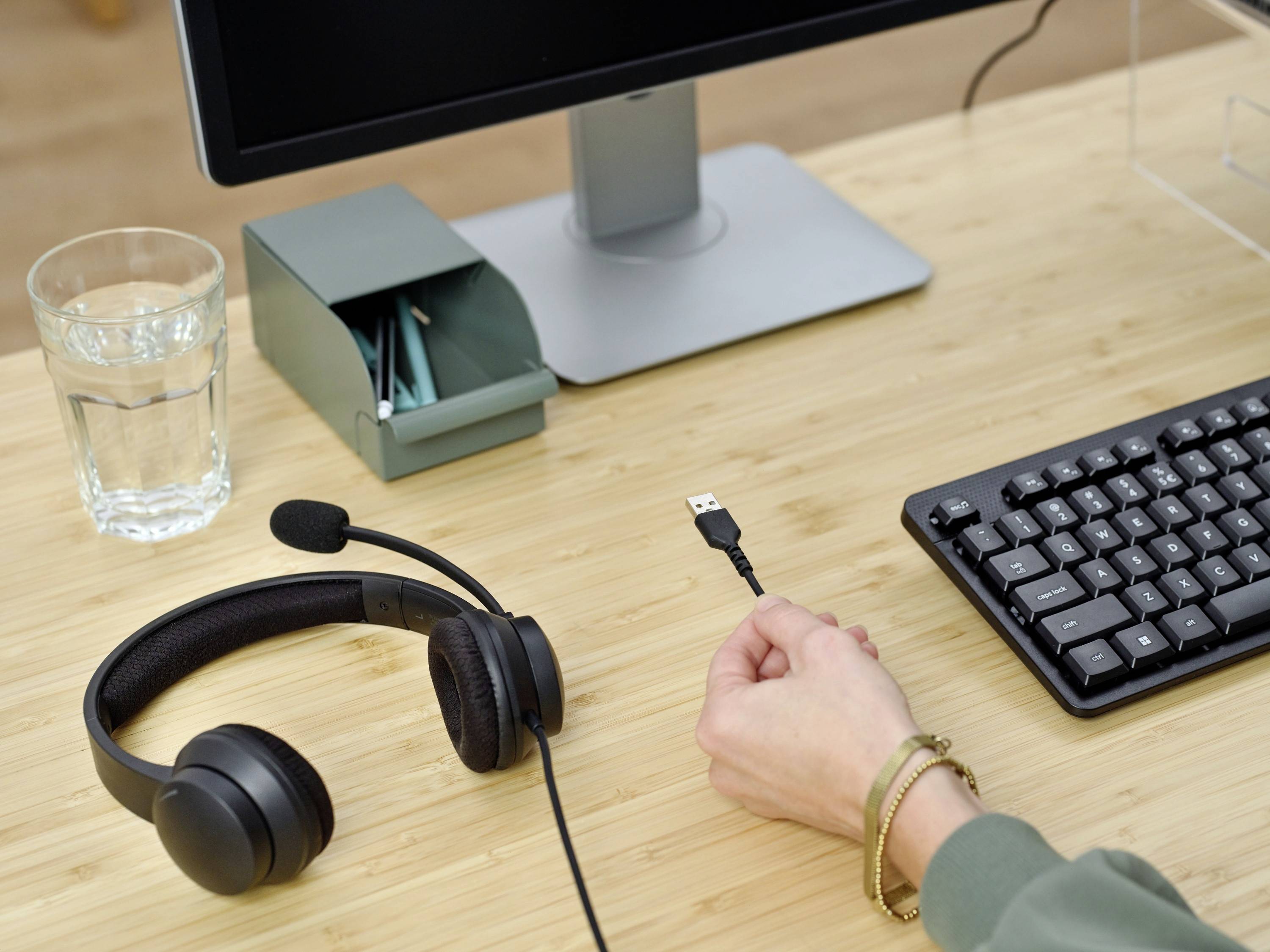 Eine Hand hält ein USB-Kabel vor einer Tastatur. Auf dem Tisch liegen ein Headset, ein Wasserglas und Stifte neben einem Bildschirm.