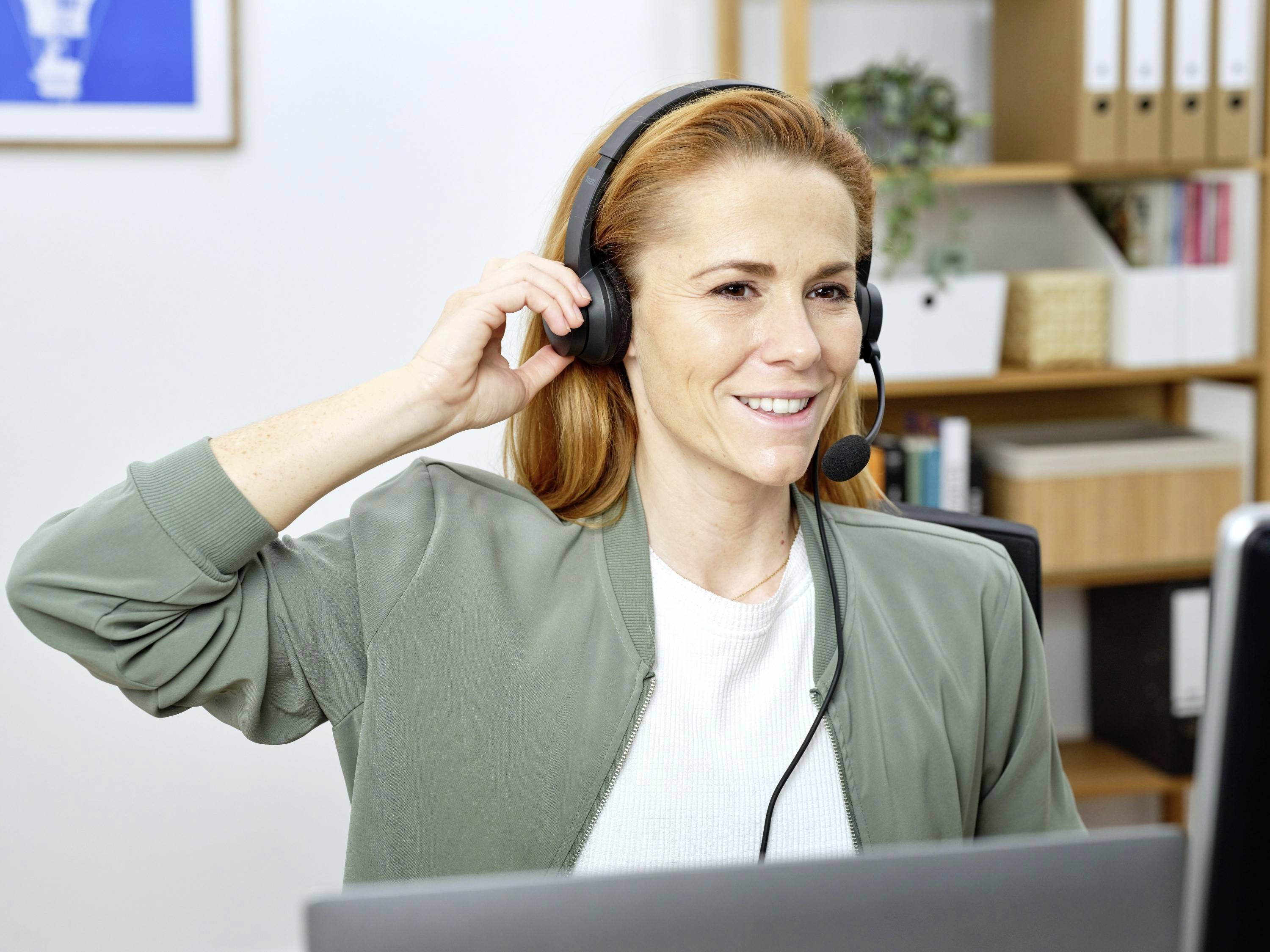 Eine Frau mit roten Haaren spricht lächelnd in einem Headset. Sie sitzt an einem Schreibtisch mit Laptop und Bücherregal im Hintergrund.