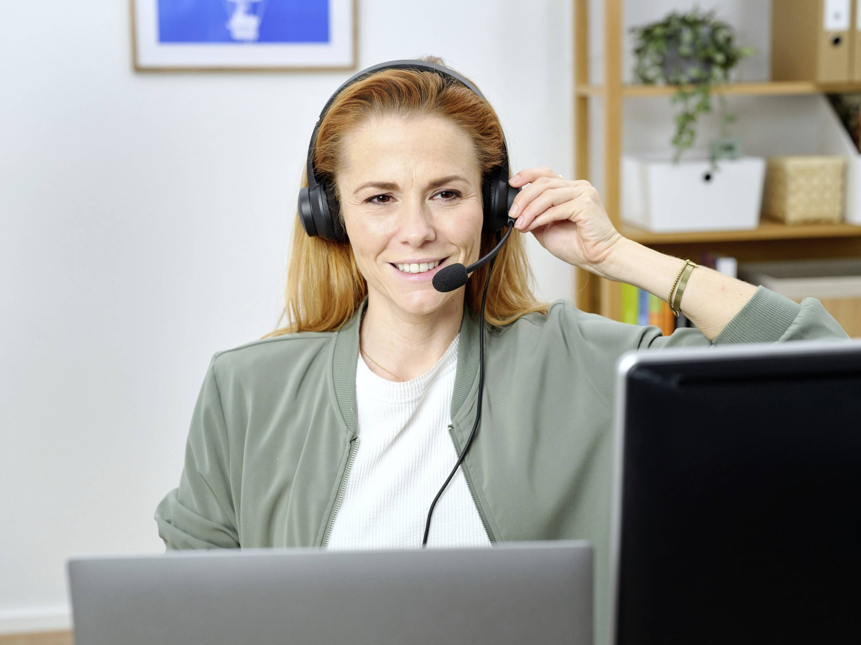 Frau mit roten Haaren sitzt vor einem Computer, trägt ein Headset und lächelt. Im Hintergrund sind ein Regal und ein Bild zu sehen.