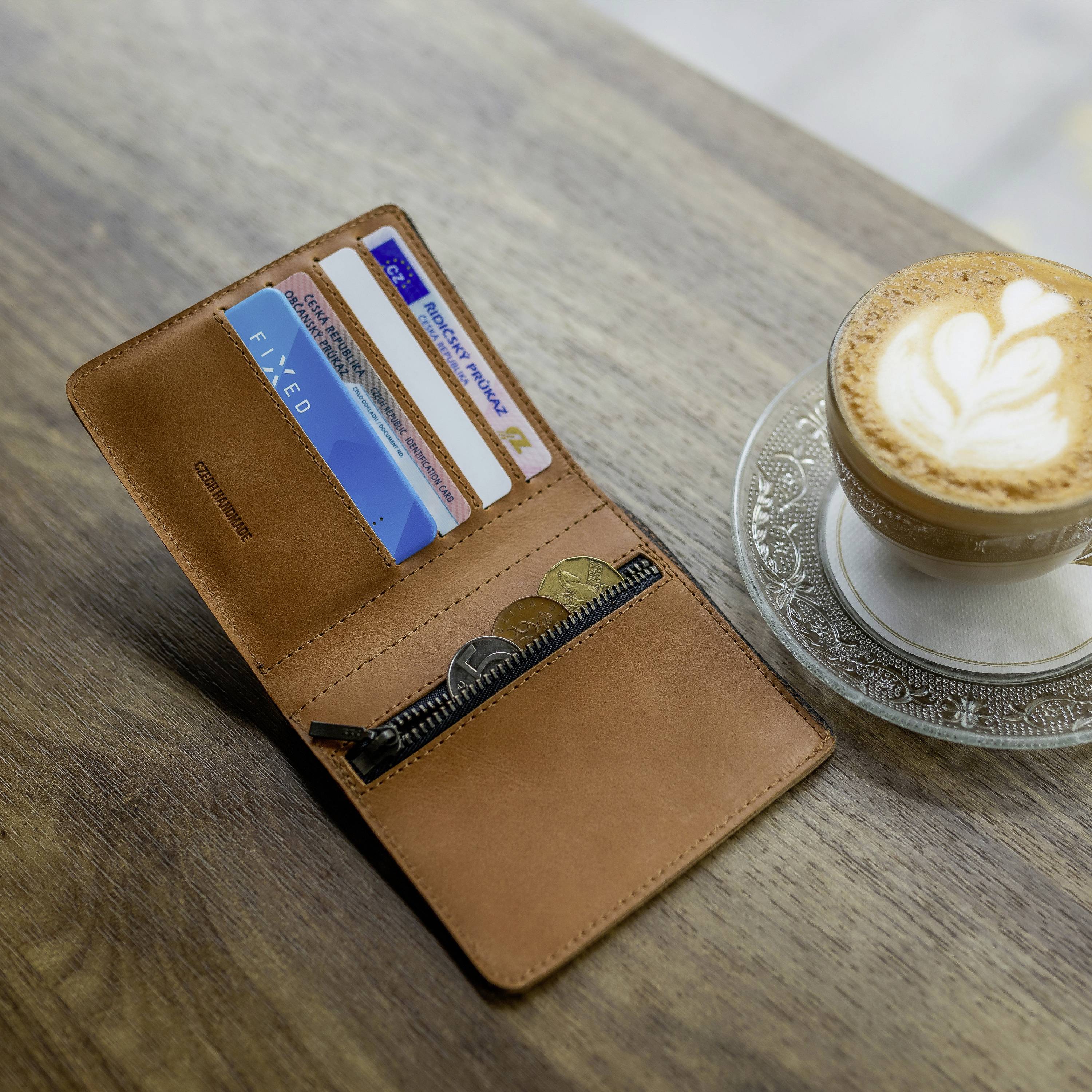 Eine geöffnete braune Geldbörse mit Kreditkarten und Münzen neben einer Tasse Cappuccino auf einem Holztisch.