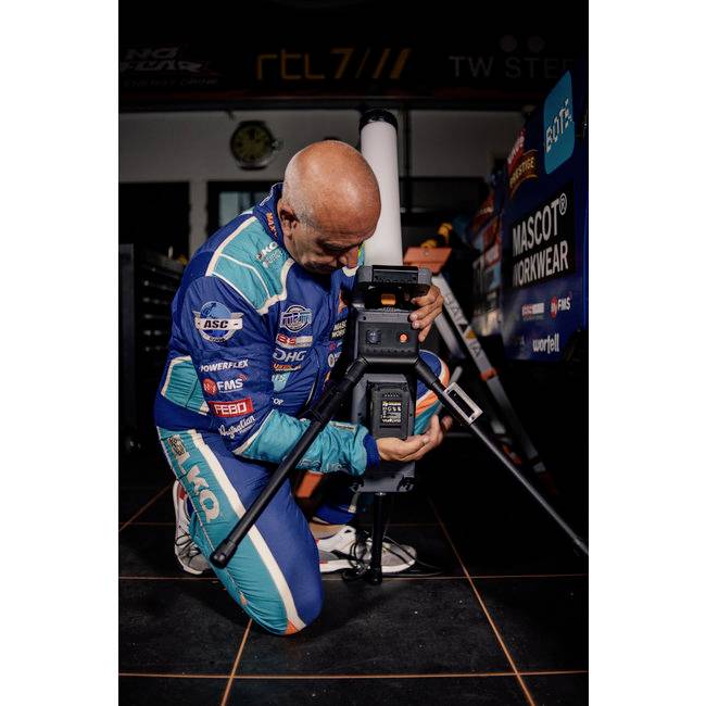 Eine Person in blauer Rennanzug kniet neben einer Kamera in einer Werkstatt. Im Hintergrund sind Sponsorlogos erkennbar.