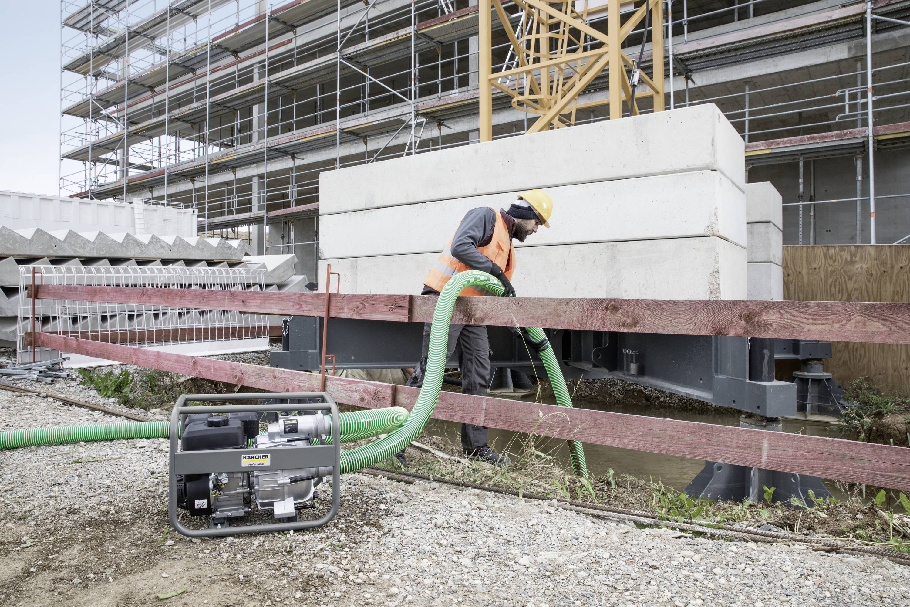 Ein Bauarbeiter pumpt Wasser auf einer Baustelle ab. Im Hintergrund sind Gerüste und Betonblöcke zu sehen.