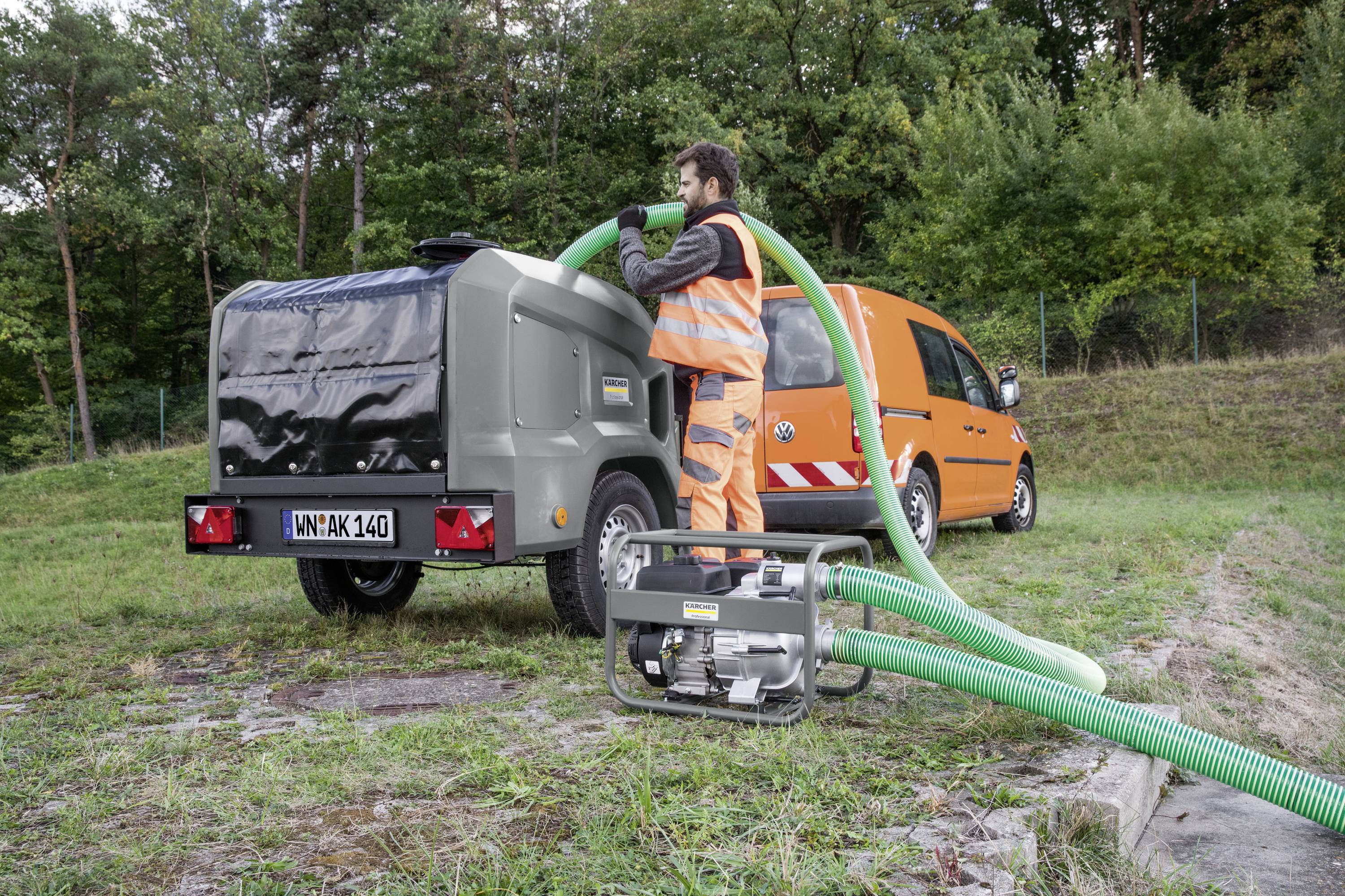 Ein Arbeiter in orangefarbener Sicherheitskleidung bedient ein Schlauchsystem an einem Anhänger neben einem orangefarbenen Firmenfahrzeug im Freien.