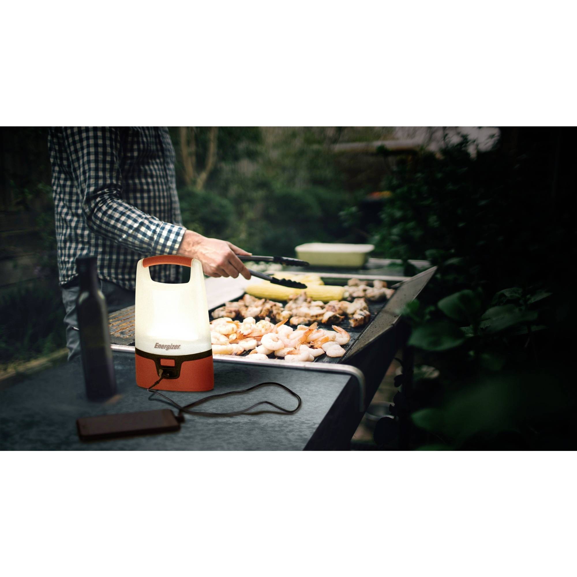 Eine Person grillt Hühnchen und Gemüse im Freien bei Dämmerung. Eine Laterne beleuchtet den Grillbereich.