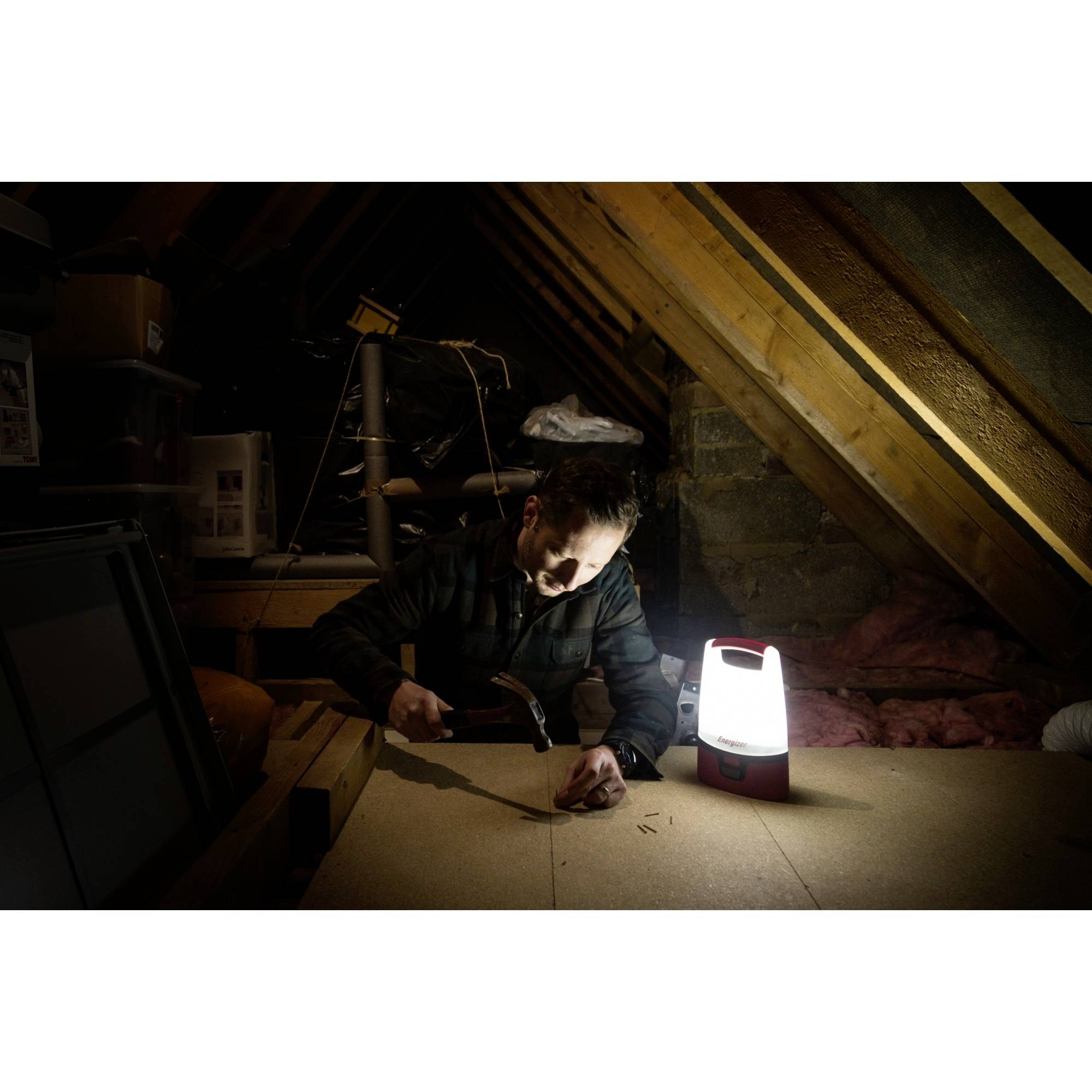 Ein Mann arbeitet mit einem Hammer auf einem Dachboden, beleuchtet von einer Lampe. Er konzentriert sich auf das Projekt vor ihm.