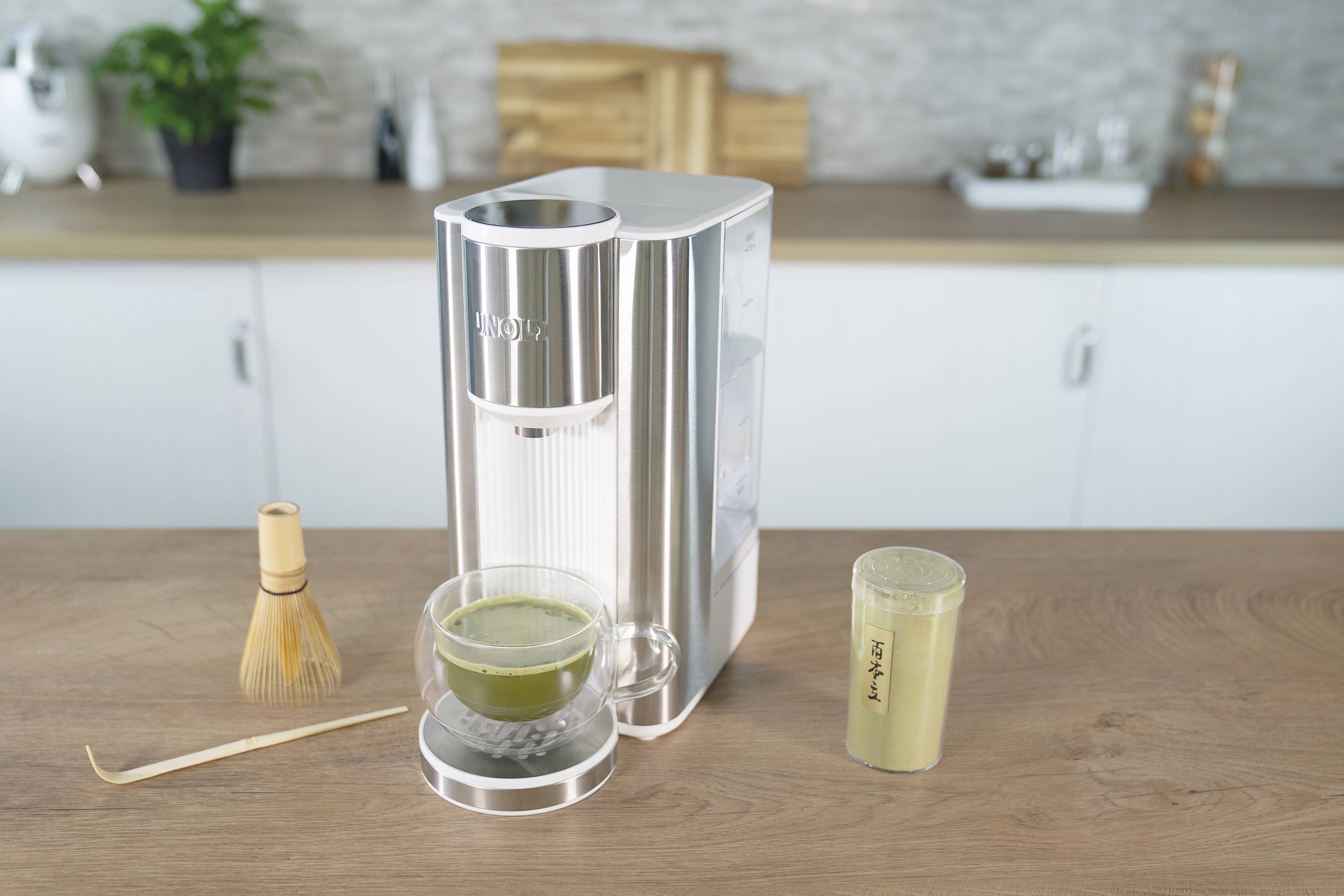 Eine Kaffeemaschine auf einer Küchenarbeitsplatte, daneben eine Tasse mit Matcha und ein Bambusbesen.
