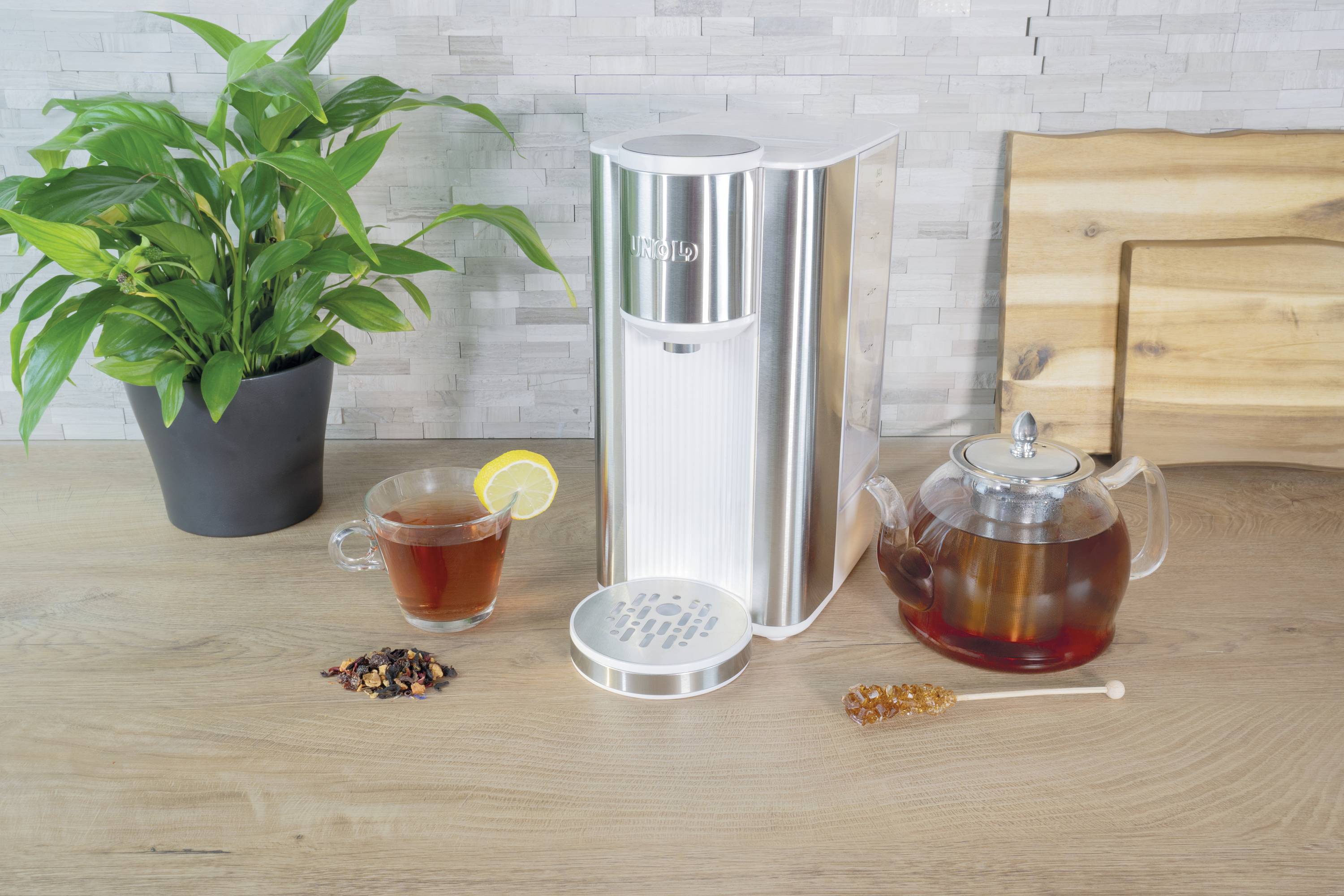 Ein silberner Wassersprudler auf einer Küchenarbeitsfläche neben einer Tasse Tee mit Zitrone, einem Teekessel und einer Pflanze.