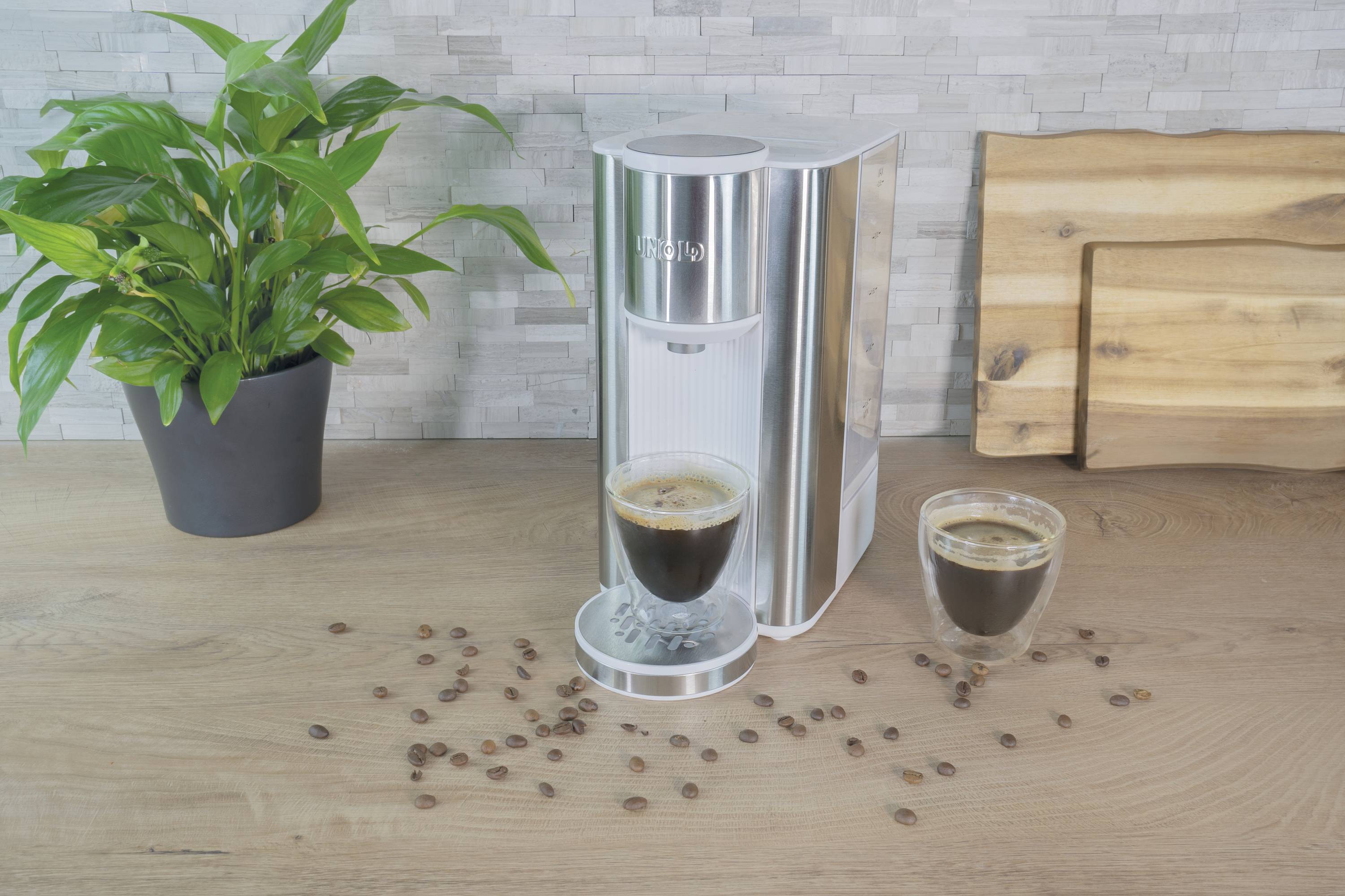 Eine silberne Kaffeemaschine auf einem Holztisch neben einer grünen Pflanze. Daneben eine Tasse gefüllter Kaffee und verstreute Bohnen.