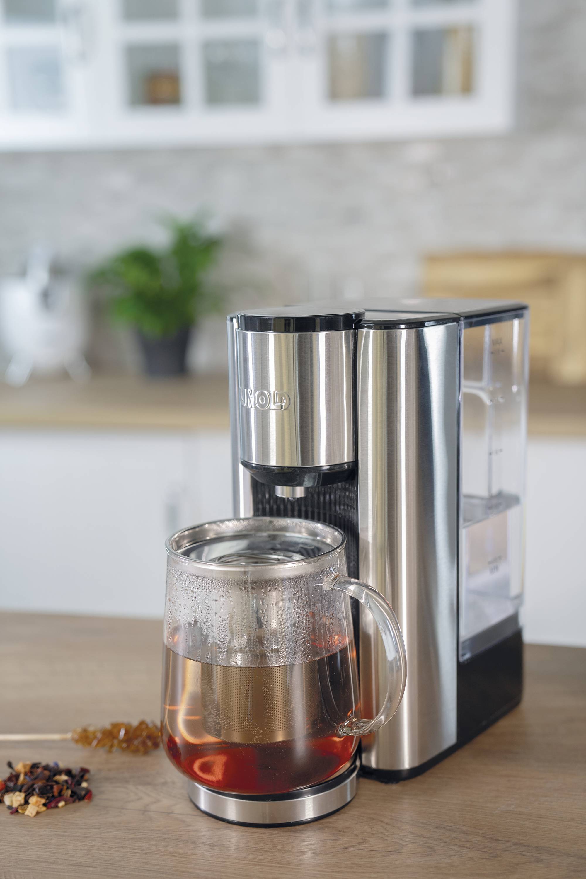 Teezubereiter aus Edelstahl auf einer Küchenarbeitsfläche, daneben eine Glasteekanne mit frisch gebrühtem Tee.