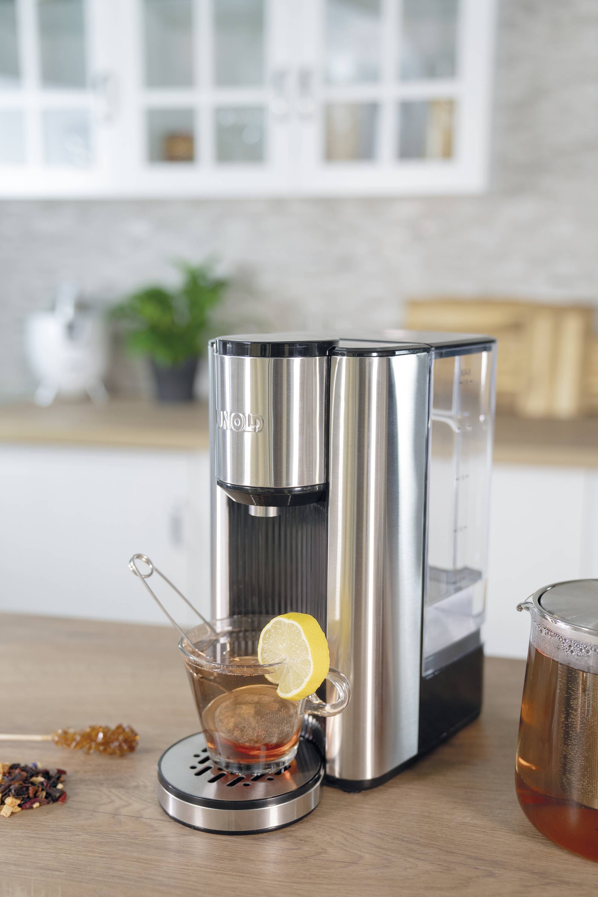 Kaffeemaschine aus Edelstahl auf Küchenarbeitsplatte. Daneben eine Glastasse mit Tee, Zitronenscheibe und Teelöffel. Küchenhintergrund.