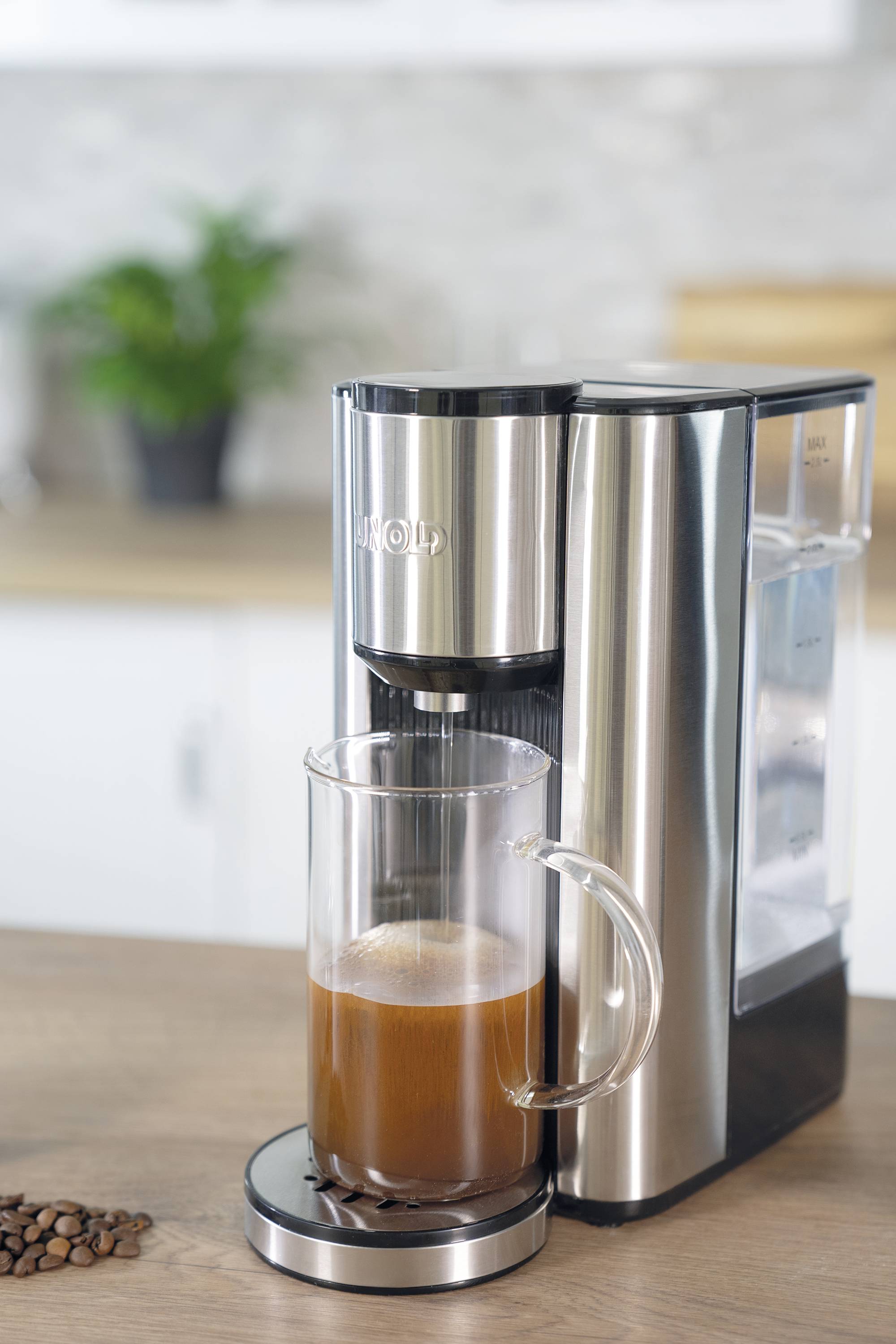 Eine silberne Kaffeemaschine auf einer Küchenarbeitsplatte brüht eine Tasse Kaffee. Im Hintergrund sind unscharfe Küchenelemente sichtbar.